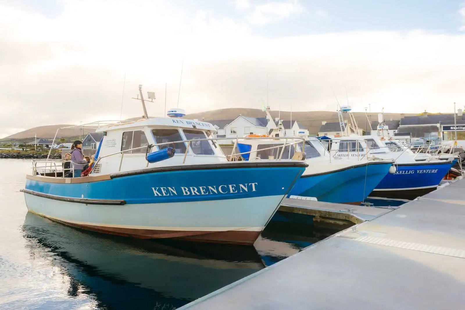 Several blue and white boats docked, preparing for Skellig Michael tours from Portmagee. Calm water reflects the vessels. 