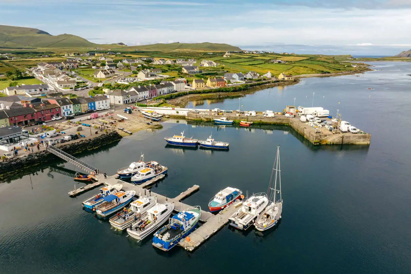 Aerial view of Portmagee harbour with boats preparing for Skellig Michael boat tours. Colourful buildings line the waterfront. Rolling hills in the distance.