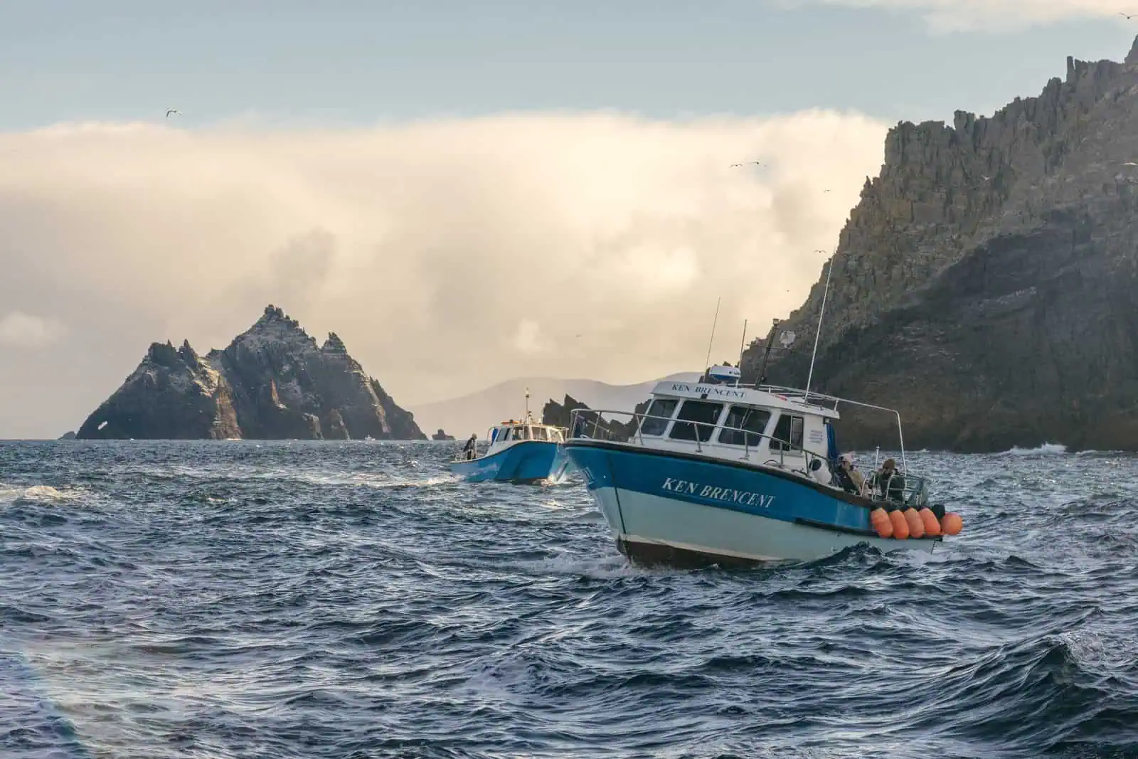 Two boats approach the rugged Skellig Islands on choppy seas, advertising Skellig Michael boat tours. Dramatic Irish coastline. 
