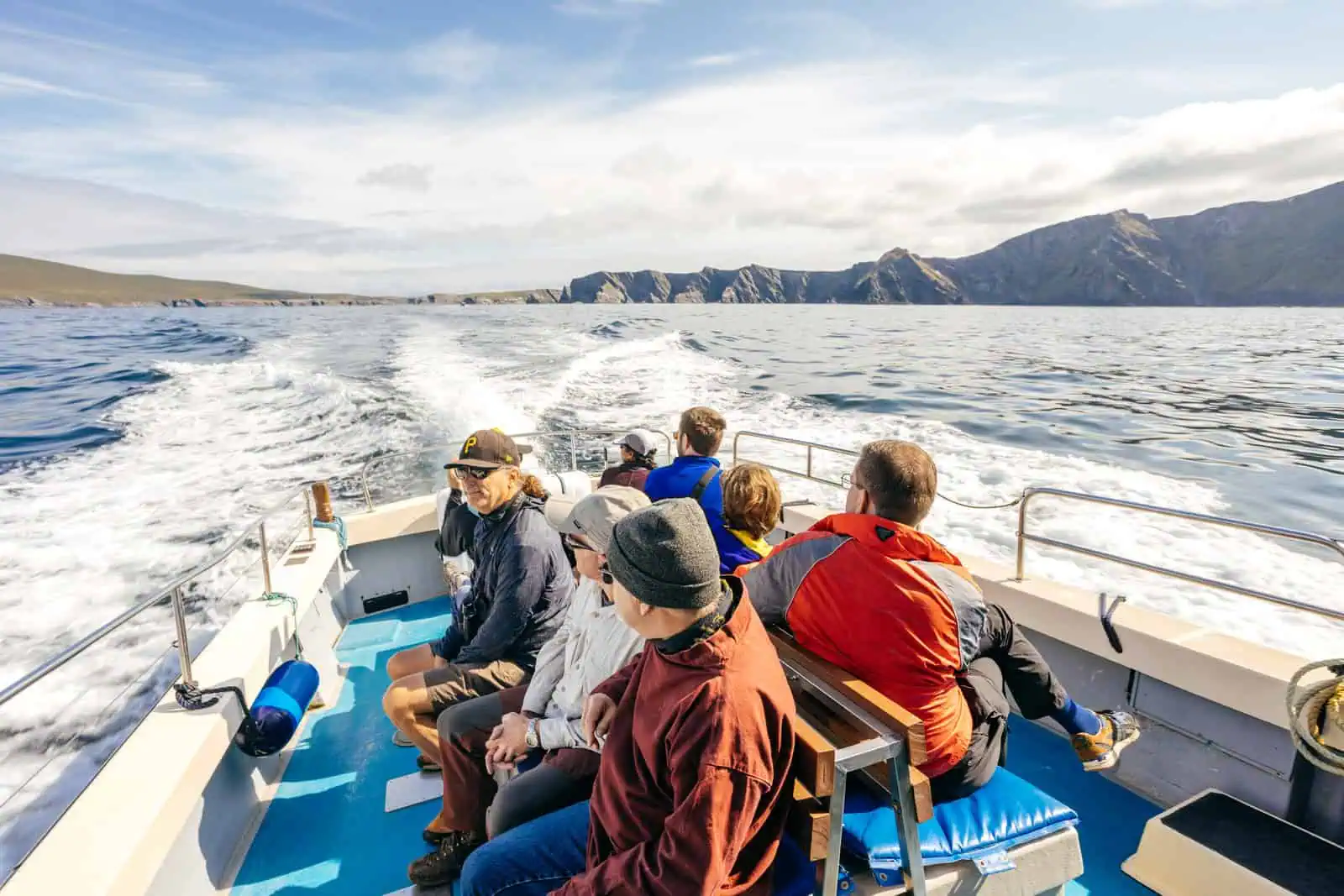 Passengers on a Skellig Michael boat tour, view from the stern towards the Skellig Islands under a bright sky. Waves visible. 