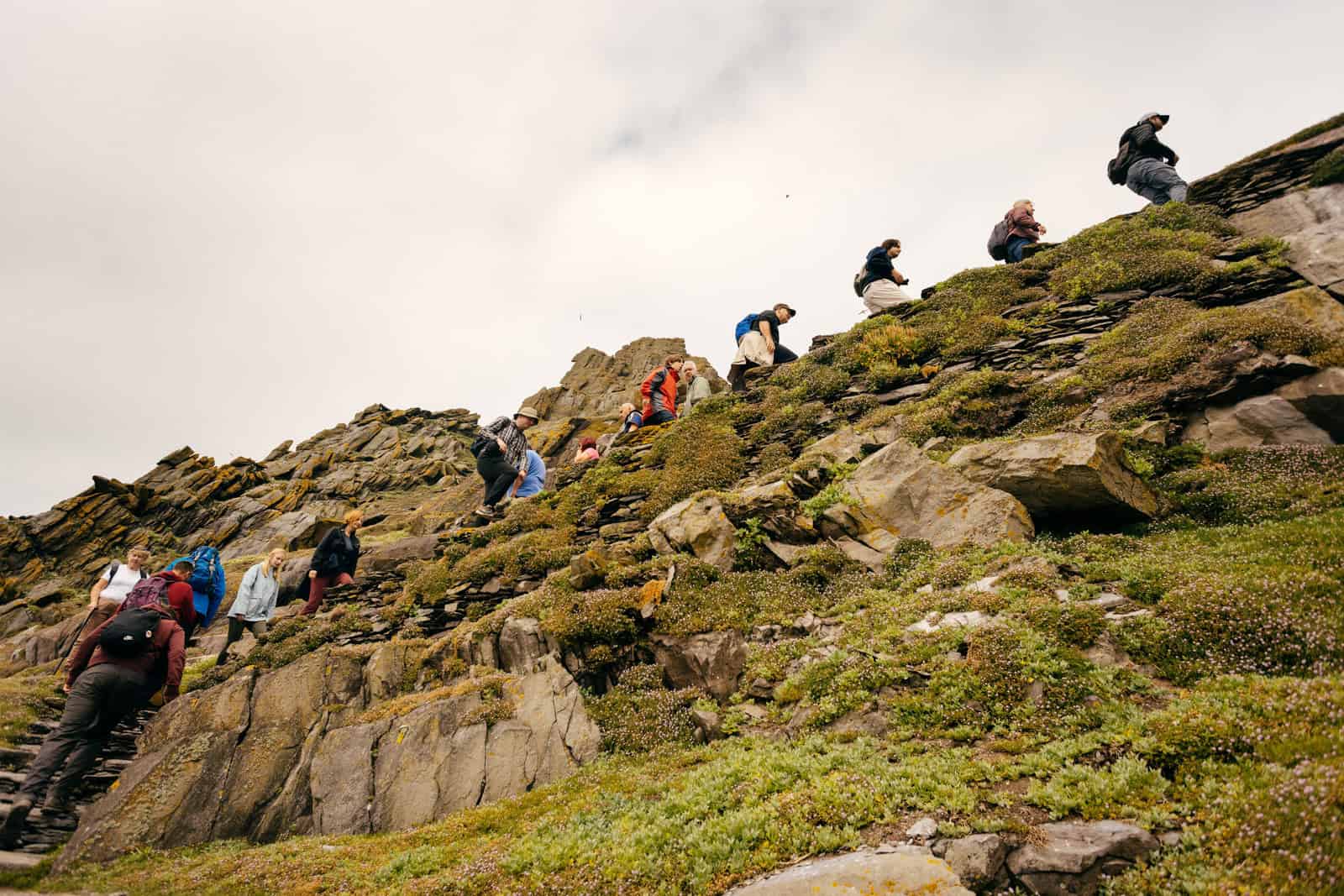 Hikers ascend a steep, rocky path on Skellig Michael. The terrain is rugged, with green vegetation clinging to the rocks. Overcast sky. 