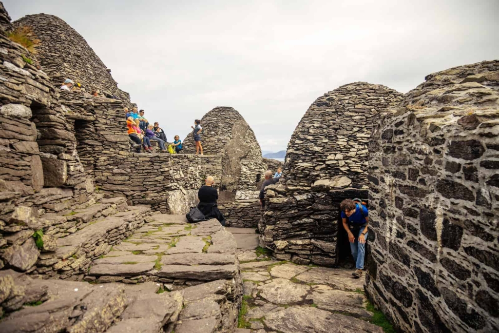 Stone beehive huts and tourists on Skellig Michael, part of the Skellig Islands Tours. Rocky terrain and cloudy sky visible.