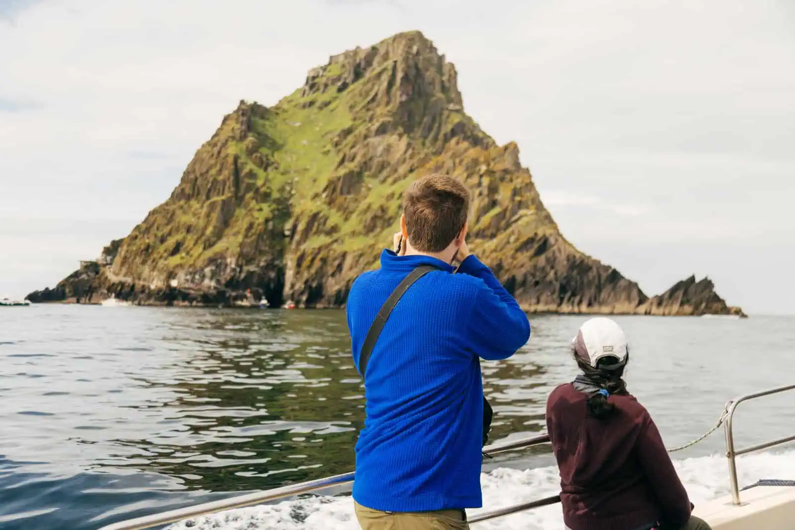 Tourists on a Skellig Michael boat tours trip, photographing the rugged, green-topped island from the sea. Dramatic cliffs visible. 