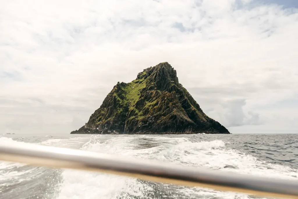 Dramatic view of Skellig Michael from a boat, offering Skellig Michael boat tours. Rocky island with green vegetation under a cloudy sky.