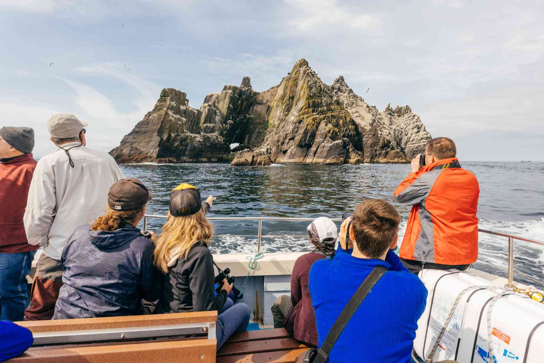 Passengers on a Skellig Michael boat tour view the rocky Skellig Islands, some pointing and taking photographs. Birds are visible.